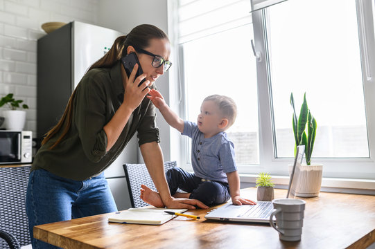 Freelancer Mother Trying To Work From Home While Babysitting With Kid. Mom Is Talking On The Phone While Her Child Is Take It Out Of Hand.