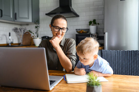 Business Mom Works From Home. Young Woman Working On A Distance With Laptop In The Kitchen, A Little Son Is Near.