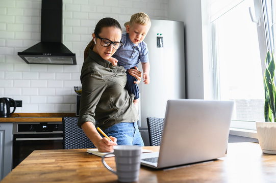 Busy mom works at home with an active toddler on her arms. Mother in glasses is watching on laptop and writing notes in the kitchen while holding kid