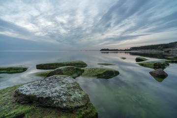 Southern coast of the Finnish Gulf. Rocks covered with green seaweed in the Baltic sea. Smooth transparent reflective water. Blue hour after sunset. Clouds concentrating in the middle. Estonia, Baltic