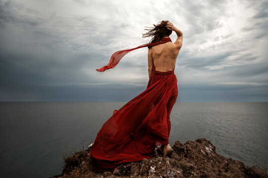 Back view a beautiful sensual woman in a red long dress poses on a rock high above the sea during sunset. Dark clouds all over the sky, a strip of fabric flying in the air.