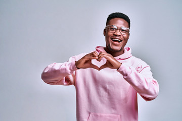 With love. Positive and curly afro man folded his hands in a heart shape, while standing against grey background
