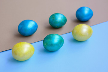 Painted yellow, blue, and green eggs lie diagonally on light blue and gold paper. Easter.