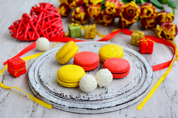 Multi-colored macaroons, yellow and red ribbons, coconut cookies and tulips on a light wooden background.