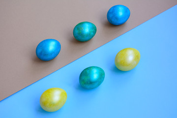 Bright blue, yellow and green eggs lie diagonally on light blue and gold paper. View from above. Holiday concept.