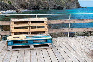 Standard wooden furniture made of cargo pallets, painted in places in blue color on the beach on a wooden platform with railings. Rocks are visible in the sea. Front view.