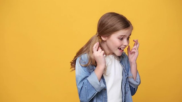 Little blonde kid teen girl 12-13 years old in denim jacket white t-shirt posing isolated on yellow background studio. People childhood lifestyle concept wait for special moment keep fingers crossed