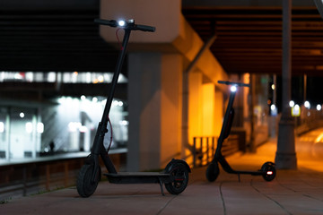 Black Electric Kick Scooters With Turn On Headlamps at Cityscape at Night Time. Scooters Under Illuminated Bridge And Near Metro Station. Urban Transportation