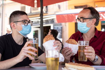 padre e figlio con mascherina chirurgica sono seduti al bar in attesa di mangiarsi un panino con la cotoletta emersi una birra 
