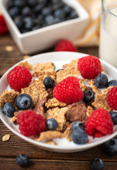 Cornflakes with blueberries and raspberries in white bowl on dark wooden desk.