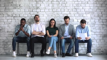 Serious young mixed race job applicants feeling nervous before interview, sitting in queue line. Thoughtful millennial multiracial diverse seekers getting bores, waiting for meeting with hr manager.