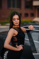 Fototapeta na wymiar Stylish young girl stands near the car in a black dress. Business fashion and style