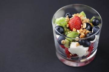 Fruit dessert made of strawberries, blueberries, kiwi, cereal and whipped cream into glass macro close up on grey / gray background