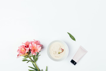 Cosmetic clay mask for skin care next to tube of cream and fresh flowers