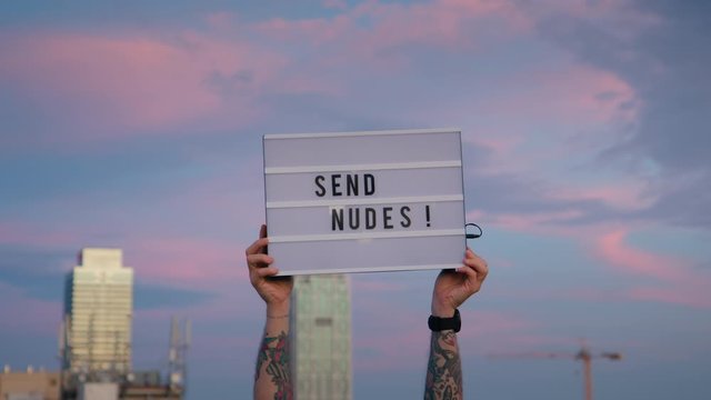 Funny sign in sky say send nudes, modern memes concept. Man holds up in city sunset sky lit up sign with light that says funny conceptual text, send nudes. Millennial era of internet jokes