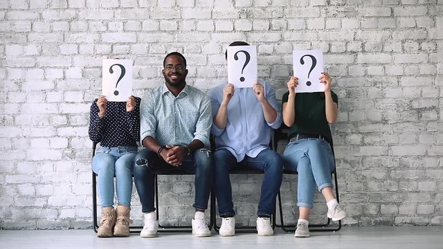 Smiling skilled young african american job applicant sitting on chair in line among unknown candidates hiding faces behinds paper sheets with questions marks, human resources, employment concept.