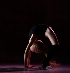 Young girl gymnast in black sport body and special footwear standing in bridge pose and keeping balance over dark background