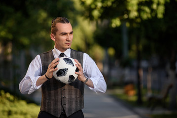 Young man football player in elegant vest, shirt and tie are holding a football ball