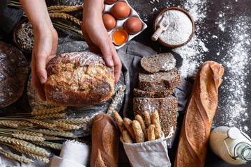 Concept of homemade bread, natural farm products, domestic production. Healthy and tasty organic food. Woman baked round whole grain bread. Top view flat lay, dark black background.