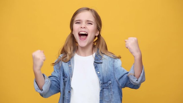 Little fun happy blonde kid teen girl 12-13 years old in denim jacket white t-shirt posing clenching fists doing winner gesture say Yes isolated on yellow background studio. People lifestyle concept