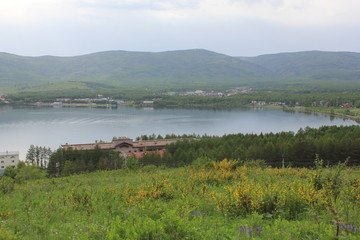 Fototapeta premium Panoramic view. A lagoon with a clean mountain lake in the midst of majestic mountains in a haze of fog. Green grass. Apartments around the lake