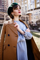 A young smiling happy girl with red lips in dark green beret and blue dress with red coat on the street. Happiness. Spring sunny day in the city. 