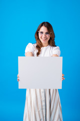 Happy young brunette woman girl in summer clothes toothy smile isolated on blue background studio portrait.Holding white big sign board blank paper overhead. Dentistry, shopping discount sale