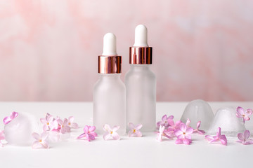 Fototapeta na wymiar Transparent cosmetic dropper bottles with ice cubes and lilac petals on pink background. Clear beauty products package without label, packaging branding mockup
