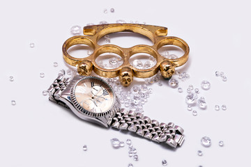 Brass knuckles and golden watch between crystal diamonds on white background, closeup with selective focus. The concept of robbery and mafia. Brass knuckle-duster, weapon for hand, isolated on white