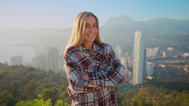 Cute and pretty, young beautiful woman or millennial zoomer teenager stand on top of hill over epic cinematic panorama of big city. Woman laugh and smily shily look at camera on sunny day