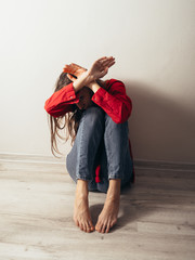 A girl in a red shirt and jeans sits sad against the wall. Domestic violence.