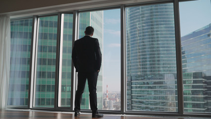 Successful businessman stands by the panoramic window. Back View of the Thoughtful Businessman wearing a Suit Standing in His Office, Hands in Pockets Looking out of the Window.