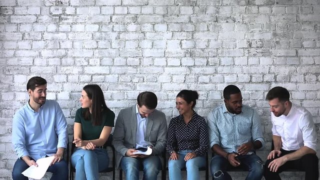 Overjoyed millennial diverse job seekers having fun, enjoying communicating in queue, waiting for interview. Happy smiling mixed race applicants chatting together, sitting in row line near empty wall.