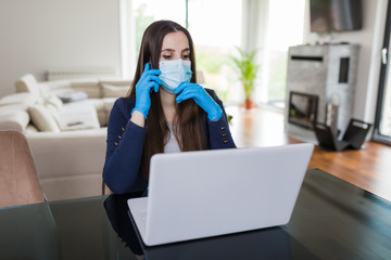 Young business woman working from home on laptop and using smartphone, wearing protective mask and gloves. Coronavirus quarantine concept.