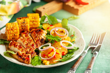  Grilled chicken breasts cooked on a BBQ served with citrus salad and corn on a plate. Close up view. Concept homemade summer dinner