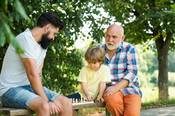 Grandfather and grandson playing chess with handsome man. Happy loving family. Grandfather and grandson are playing chess and smiling while spending time together in park.