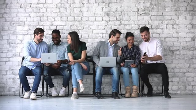Happy communicative millennial diverse people sitting on chairs, sharing projects experience on computers with each other. Smiling sociable applicants having fun, waiting for job interview in queue.