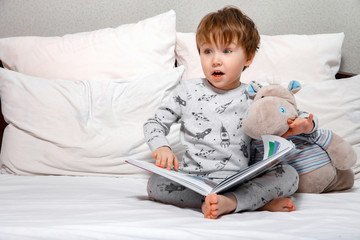 Little cute boy in pajamas reading a toy book sitting on the bed. Bedtime stories before bed.