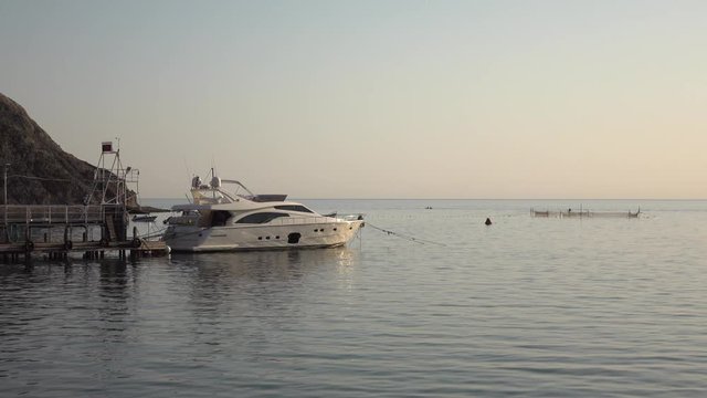 Luxury white sea yacht moored in solitary marina at sunset. Sea is calm, without waves. Rays of the pre-sunset sun are reflected on smooth surface of water. Skyline and mountains are visible