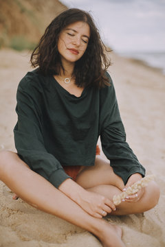 Stylish hipster girl holding herb and sitting on sandy beach. Fashionable tanned young woman in modern swimsuit and sweater relaxing on seashore. Summer vacation. Carefree