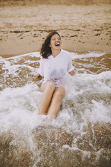 Fototapeta na wymiar Happy young woman in wet white shirt lying on beach in splashing waves. Emotional tanned girl relaxing on seashore and enjoying waves. Summer vacation. Carefree moment