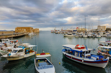 Fototapeta na wymiar Heraklion, Crete / Greece. Panoramic view at the old Venetian port. Traditional and colorful fishing boats are docked. Venetian fortress Koules in the background. Sunset with cloudy sky