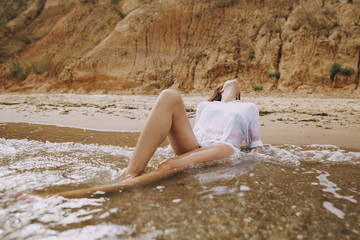 Young woman in white shirt sitting on beach in splashing waves. Stylish tanned girl relaxing on seashore and enjoying waves. Summer vacation. Mindfulness and carefree moment