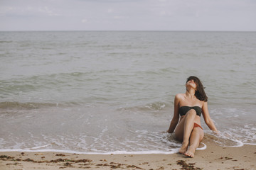 Fototapeta na wymiar Happy young woman sitting on beach with waves. Stylish tanned girl in modern swimsuit relaxing on seashore. Summer vacation. Carefree moment. Authentic image