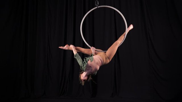 The flexible athletic circus gymnast performs acrobatic elements on the air ring. Circus performer spinning in a split on a background of black backstage scenes. young aerialist on aerial hoop.