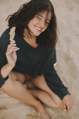 Carefree hipster girl with windy hair sitting and smiling on sandy beach, holding herb. Stylish...