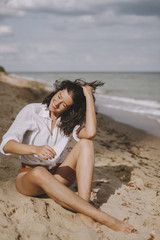 Fototapeta na wymiar Joyful boho girl in white shirt sitting on sunny beach. Carefree stylish woman smiling and relaxing on seashore. Summer vacation. Lifestyle authentic image. Mindfulness.