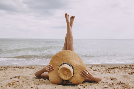 Girl in hat lying on beach with legs up. Fashionable young woman covering with straw hat, relaxing on sandy beach near sea. Summer vacation and travel. Mindfulness and carefree