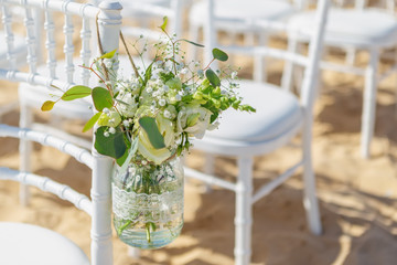 White chairs at a beach wedding ceremony, decorated with a hanging jar of flowers, on a sunny day. Close-up.