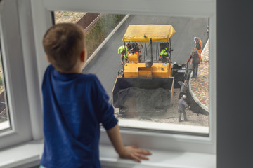 Male builders make a road near a residential complex. The boy looks out the window, where the...